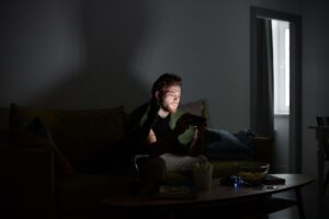 A gamer sitting in a dark room illuminated by monitor light, focused on screen